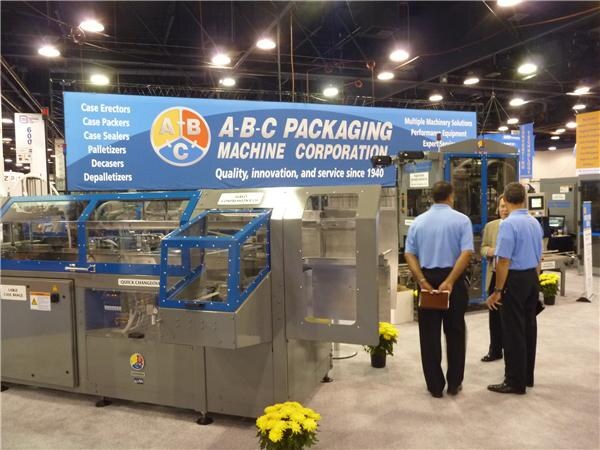 A packaging machine with blue framing sits under an A-B-C Packaging Machine Corporation sign. Two men in blue shirts stand nearby, and yellow flowers decorate the booth.