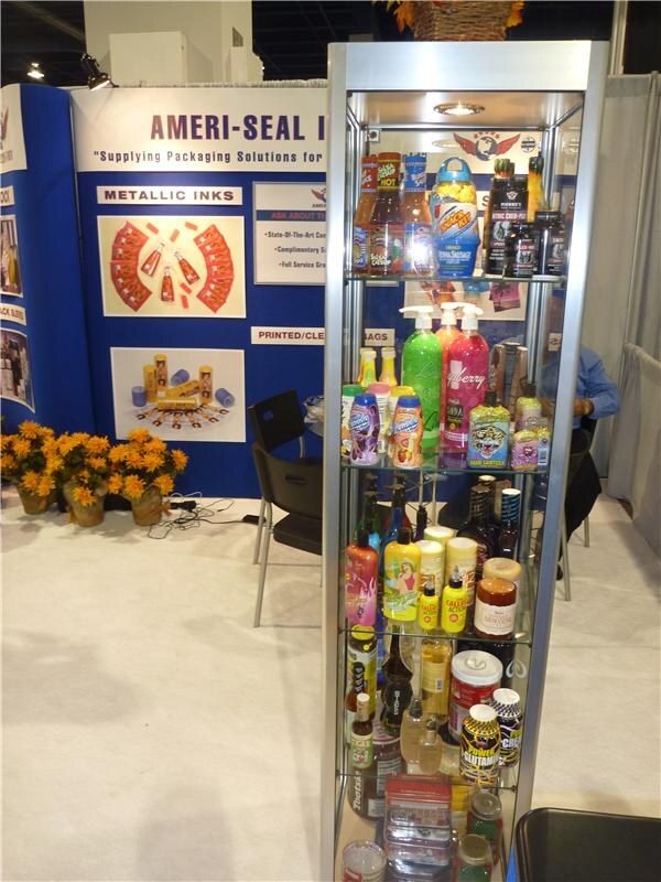 A glass display case holds bottles and containers with printed labels in front of an Ameri-Seal booth wall. The wall shows metallic inks graphics, and yellow flowers sit on the floor.