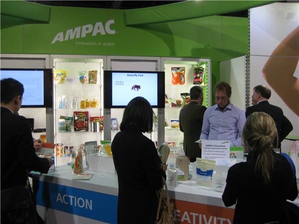 Visitors and booth staff gather at a counter beneath an Ampac sign. Shelves behind them hold pouches and packaging samples, and screens show a Butterfly Pack slide.