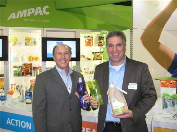Two smiling men in suit jackets stand at the Ampac booth. Each holds flexible pouches or a jug, with shelves of packaging samples and an Ampac sign behind them.