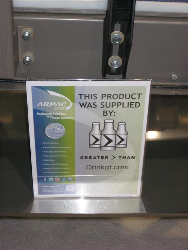 A sign beside a machine reads "This product was supplied by: Greater Than, Drinkgt.com." It also carries Arpac Group branding and a list of its packaging services.