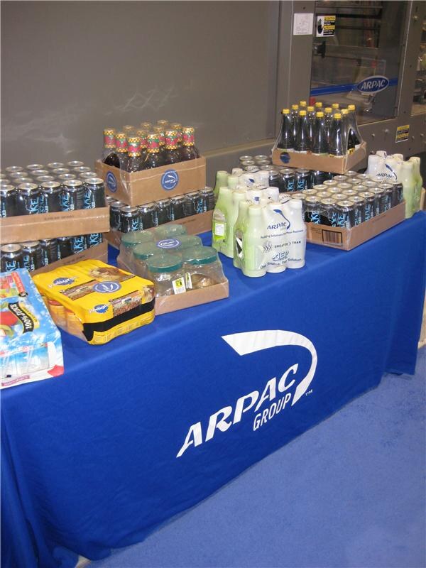 A blue table with an Arpac Group logo holds shrink-wrapped bundles of bottles, cans and jars, and a machine with an Arpac logo stands behind it.