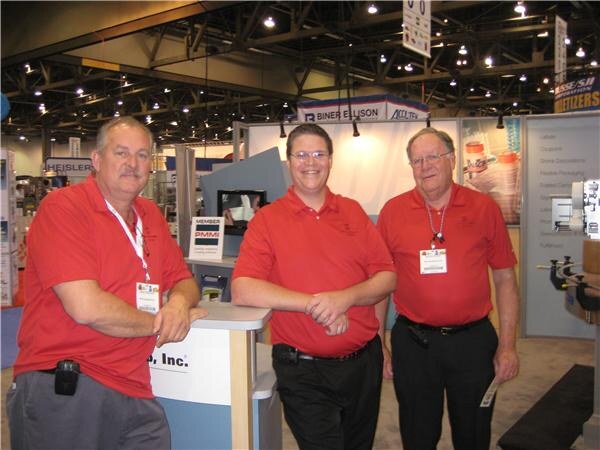 Three men in red polo shirts and badges stand at a counter in an exhibit hall, with a Biner Ellison sign and a PMMI member sign visible behind them.