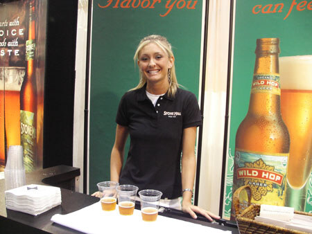 A smiling woman in a dark Stone Mill polo stands behind a counter with three small cups of beer, near a Wild Hop Lager bottle graphic and a basket of items.