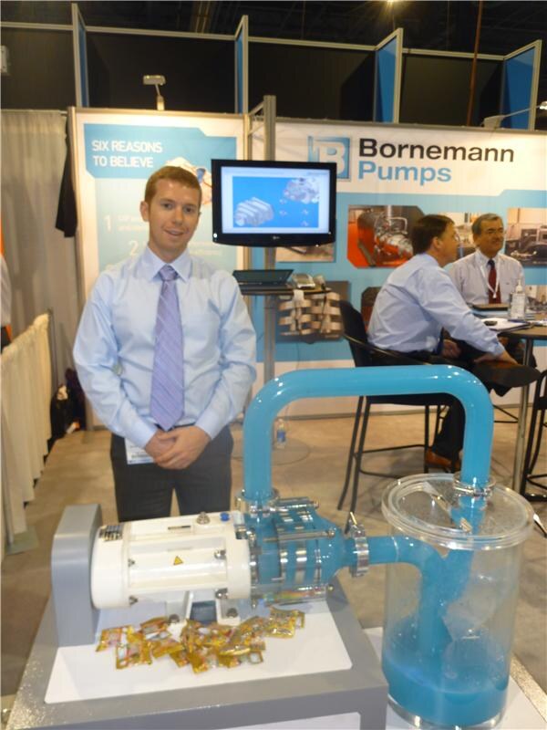 A young man in a blue shirt and tie stands behind a pump demonstration with blue liquid in a clear container. Bornemann Pumps signs and two seated men appear behind him.