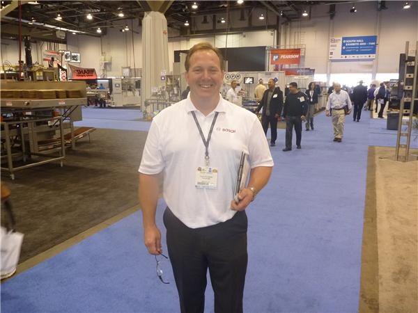 A smiling man in a white Bosch polo shirt with a lanyard and badge stands on blue carpet in an exhibit hall, holding a folder, with other attendees walking behind him.