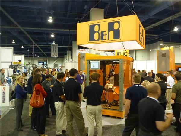 An orange B&R sign hangs over an orange automation machine in an enclosure. A crowd of attendees stands around the booth in the exhibit hall.