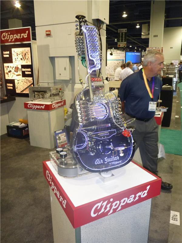 A clear acrylic guitar labeled Air Guitar by Clippard, with air valves and tubing and lit edges, sits on a Clippard pedestal. A man in a navy polo stands beside it.