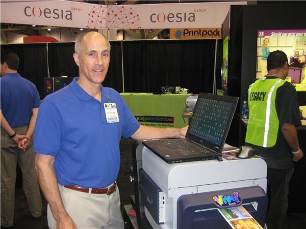 A man in a blue polo shirt with a badge stands beside a laptop on a printer in a booth under Coesia Group and Printpack signs; a man in a safety vest stands at right.
