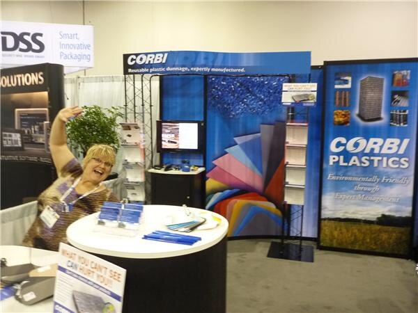 A smiling woman with blonde hair raises one arm at a round counter in the Corbi Plastics booth, which has blue banners reading Corbi Plastics and colorful plastic sheet displays.