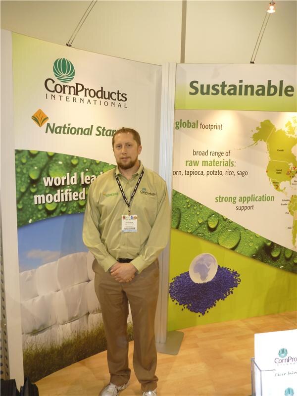 A bearded man in a green button-up shirt with a badge and lanyard stands in front of CornProducts International and National Starch banners that mention sustainability.