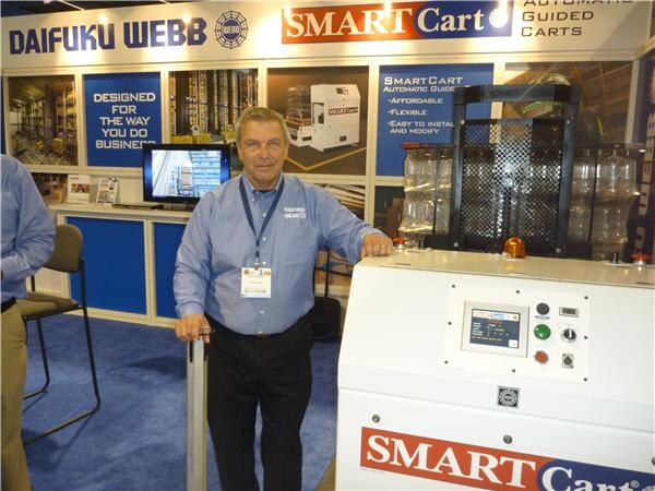 A man in a light blue shirt and lanyard stands beside a white SmartCart automatic guided cart in the Daifuku Webb booth, with SmartCart signs and a monitor behind him.