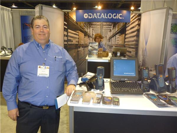 A man in a blue button-up shirt with a badge stands at a Datalogic booth table displaying handheld scanners and a rugged computer terminal, with a woman seated behind.