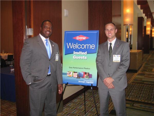 Two men in gray suits with name badges stand on either side of a Dow sign reading Welcome Invited Guests, Dow Performance Plastics, in a carpeted hall.
