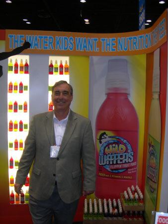 A smiling man in a gray blazer stands in front of a Wild Waters display with a large red Wild Waters bottle and a banner reading "The water kids want."
