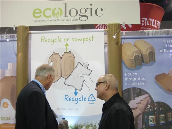 Two men look at an Ecologic banner showing a recycle or compost diagram of a paper shell and pouch, with nested shells pictured at right.