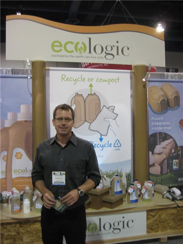 A man with glasses and a dark striped shirt stands in front of the Ecologic booth, holding a small package. Banners about recycling and composting and Ecologic bottles are behind him.