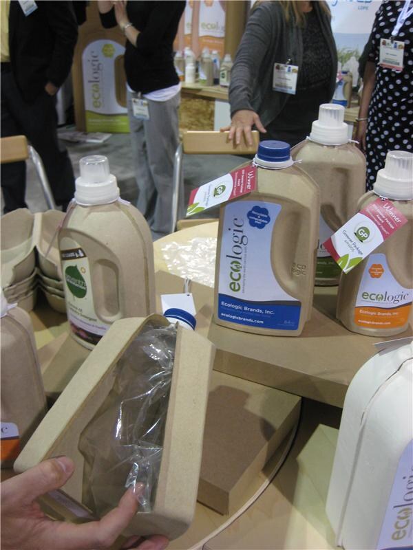 Molded paper-fiber Ecologic detergent bottles with award tags sit on a round display, while a hand holds a fiber shell with a plastic pouch and attendees stand behind.