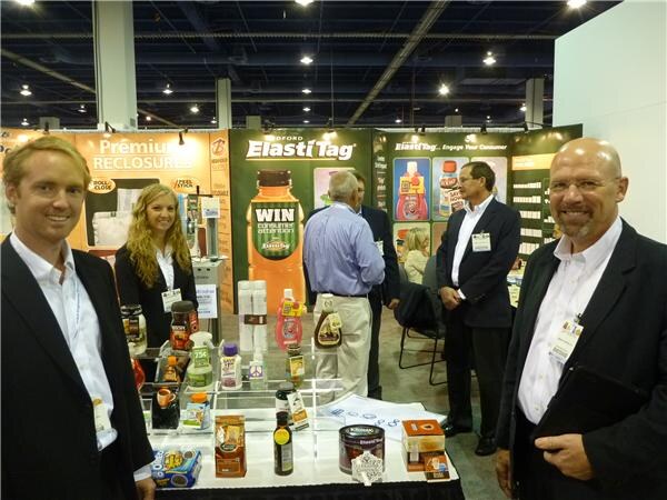 Four men and a woman stand at the ElastiTag booth, with a table of bottles and jars in front. Behind them, a sign reads ElastiTag and a bottle graphic reads WIN.
