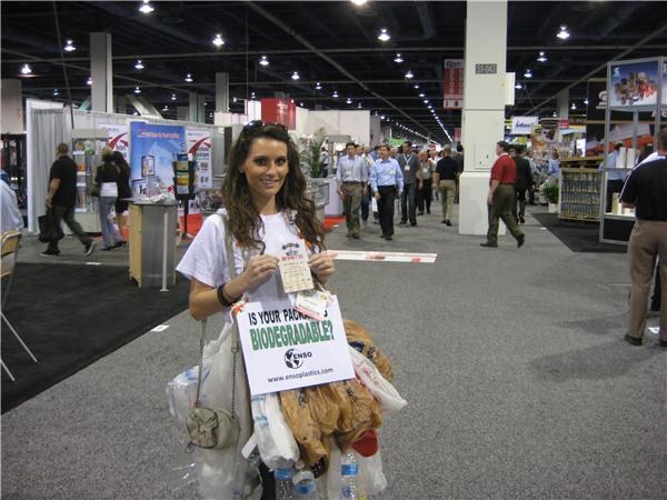 A smiling woman with long dark hair holds a sign reading Is your package biodegradable? with the Enso Plastics web address, surrounded by plastic bottles and bags on the show floor.