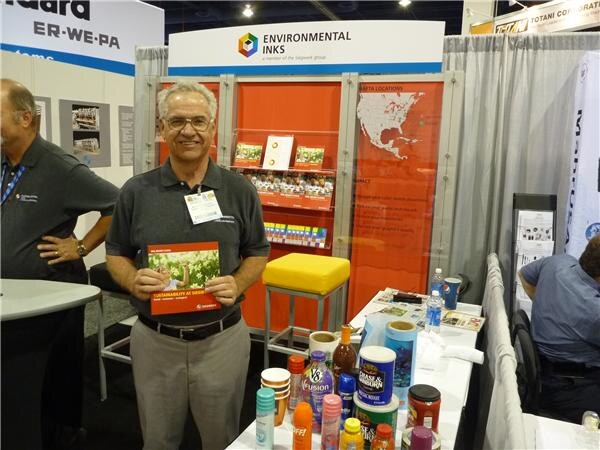 A smiling man with glasses and a gray polo holds a booklet at the Environmental Inks booth. A table of product containers is nearby, and a Totani Corporation sign hangs at the top.