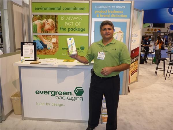 A man in a green polo holds up a carton beside the Evergreen Packaging booth counter. A sign behind reads environmental commitment is always part of the package.