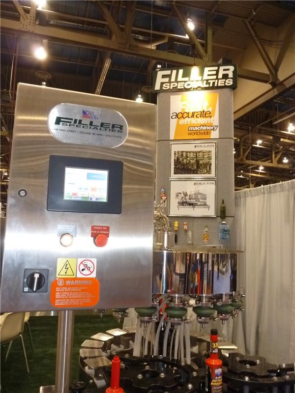 A stainless steel control panel with a touchscreen and emergency stop button stands beside a Filler Specialties filling machine with bottles, under a Filler Specialties sign.