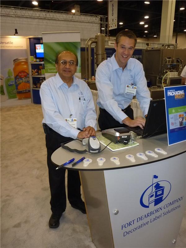 Two men in light blue shirts stand at a Fort Dearborn Company counter, one using a laptop. A Packaging Digest sign sits on the counter, and Dial and SunFresh products are visible behind.