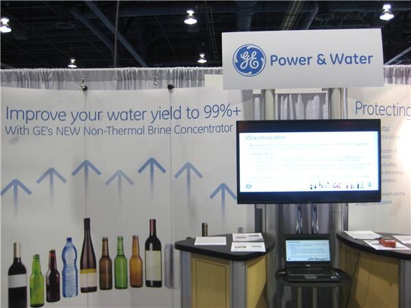 The GE Power & Water booth with a banner reading Improve your water yield to 99%+ with GE's new Non-Thermal Brine Concentrator, bottle images, a monitor and a laptop on counters.