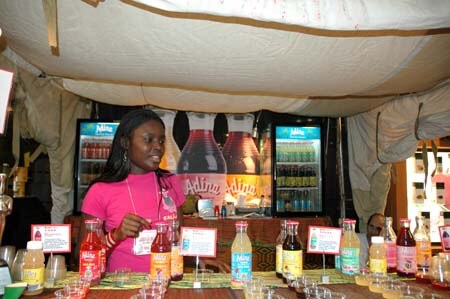 A woman in a pink shirt stands behind a table of bottled Adina drinks and small signs, with Adina branded refrigerators and large bottle graphics behind her.