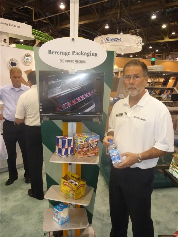 A bearded man in a white polo holds a water bottle beside a Graphic Packaging Beverage Packaging display with cartons including Hiland. A Scandia Packaging Machinery sign hangs overhead.