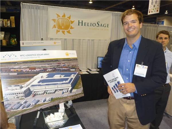 A smiling man in a navy blazer and blue shirt holds a brochure beside a HeliSage booth display of an aerial photo of the MTC Logistics solar array; another man stands behind him.