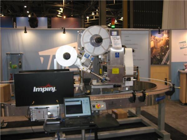 A labeling machine with two label reels over a curved conveyor, with an Impinj-branded monitor and a laptop on a stand in front, inside a trade show booth.