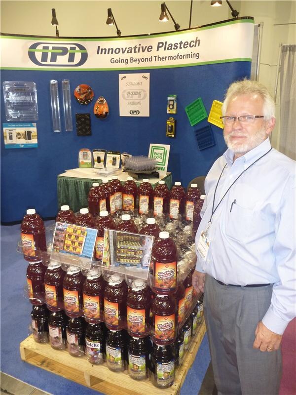 A bearded man with glasses and a lanyard stands beside a pallet of Welch's blackberry juice bottles in front of an Innovative Plastech booth showing thermoformed plastic packaging samples.