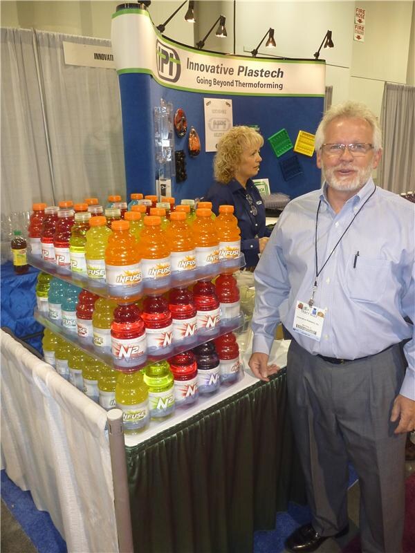 A bearded man with glasses stands beside tiered displays of colorful Infuse bottles at the Innovative Plastech booth, while a woman with curly blonde hair stands in the background.