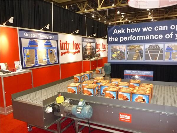 An Intralox booth with red walls and signs reading Greater Throughput, with a conveyor belt in the foreground carrying orange boxes and a large banner above.