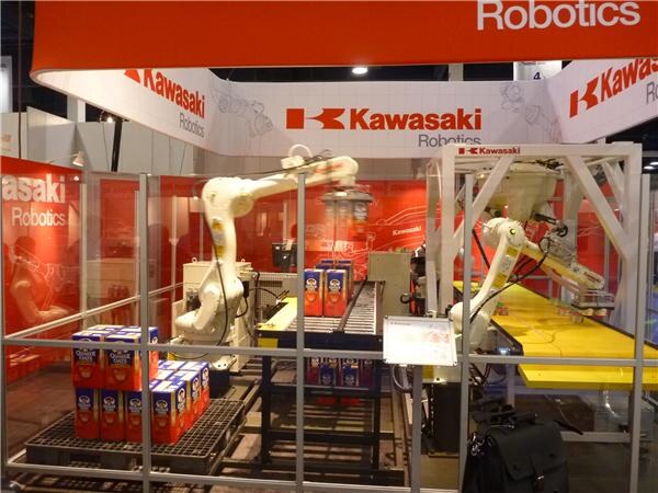 Two white Kawasaki robotic arms handle cartons of Quaker products at conveyors inside a glass-enclosed red Kawasaki Robotics booth, with stacked cartons on a pallet at left.