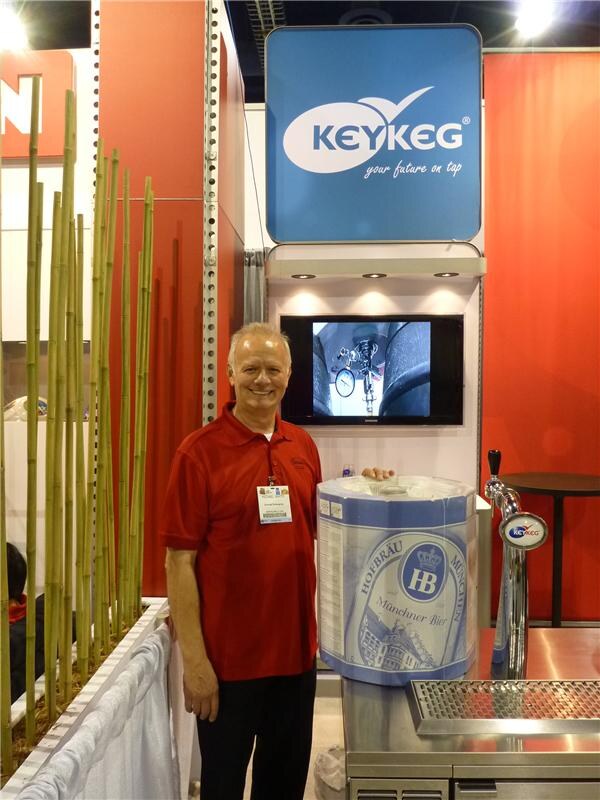A smiling man in a red polo shirt stands beside a Hofbräu München KeyKeg keg and a tap in front of a blue KeyKeg sign. A Samsung monitor hangs on the wall above, and bamboo stalks stand at left.