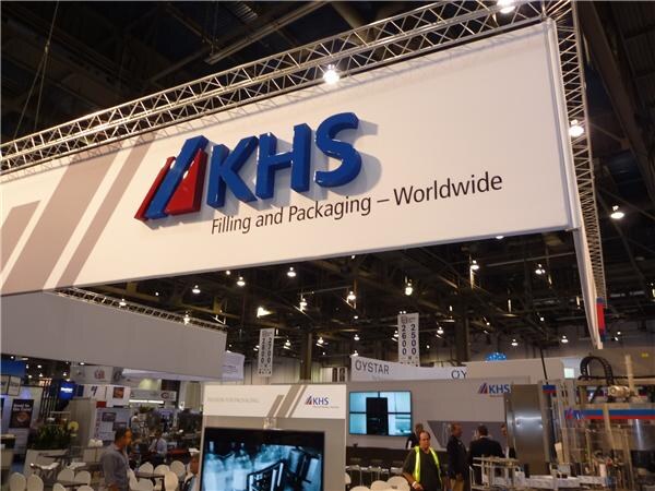 A large white KHS sign reading Filling and Packaging – Worldwide hangs from a truss over the booth, with an Oystar sign and several attendees visible in the background.