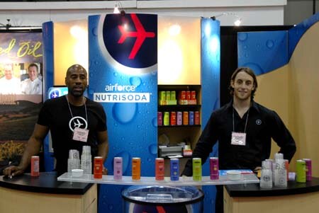 Two men in dark shirts and name badges stand at the Airforce Nutrisoda booth, with colorful cans lined up on the counter between them and a blue branded backdrop.