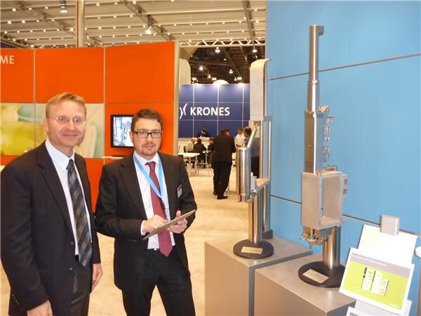 Two men in dark suits stand beside metal machine components on display stands at the Krones booth, one holding a tablet, with a Krones sign in the background.