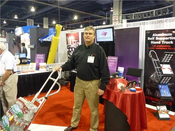 A man in a black shirt and khaki pants stands beside an aluminum hand truck loaded with water bottles in the Magnus booth, with a Magnus monitor and a hand truck banner behind him.