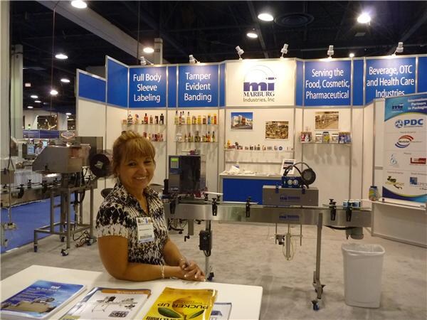 A smiling woman with a name badge sits at a table in the Marburg Industries booth, with a labeling machine behind her and signs for sleeve labeling and tamper evident banding. A PDC banner is at right.