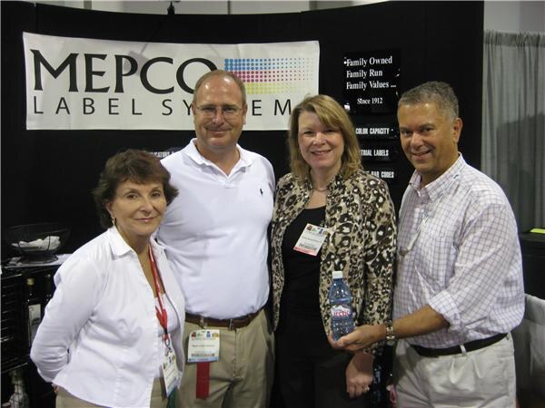 Four people, two women and two men, stand together in front of a MEPCO Label Systems banner. One woman wears a leopard-print jacket, and the man at right holds a water bottle.