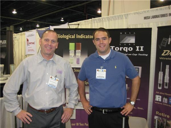 Two smiling men wearing name badges stand in front of purple Mesa Labs display banners that mention biological indicators and Torqo II cap testing.