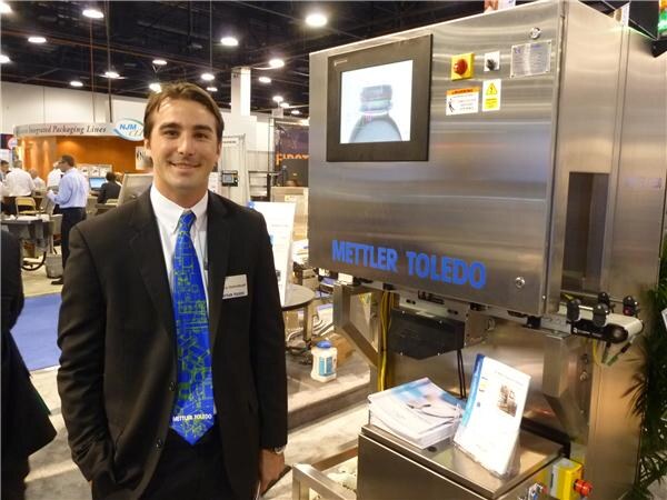 A smiling man in a dark suit and a blue patterned Mettler Toledo tie stands beside a stainless steel Mettler Toledo inspection machine with a screen showing a bottle.
