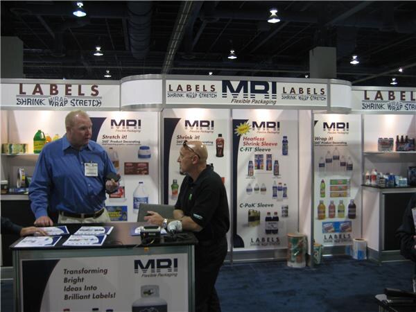 Two men talk at the MRI Flexible Packaging booth. Panels behind them show shrink sleeve, stretch and wrap labels, and sample bottles.