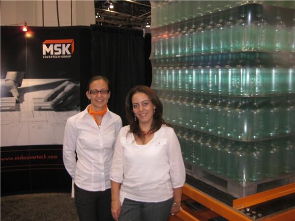 Two women in white tops stand in front of an MSK Covertech Group banner, next to a tall stack of water bottles on a pallet.
