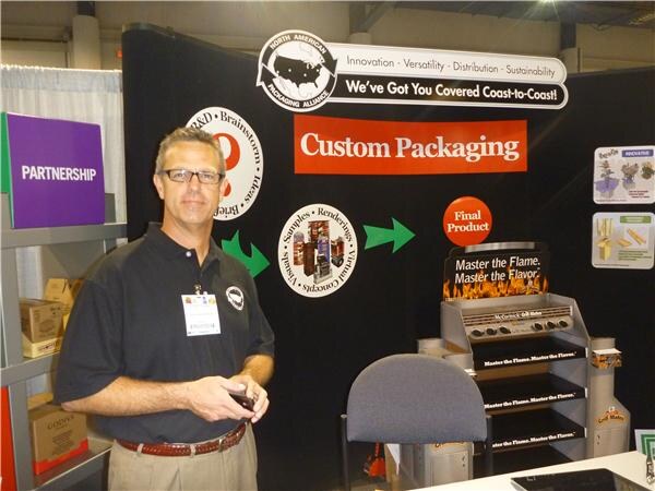 A man in glasses and a black polo stands in the North American Packaging Alliance booth, with a Custom Packaging banner and a Grill Mates display behind him.