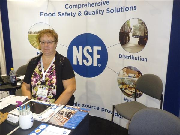 A woman with glasses and a lanyard sits at a table in front of an NSF banner reading Comprehensive Food Safety & Quality Solutions, with brochures and a cup of pens on the table.